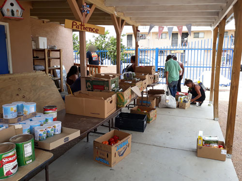 open boxes of food and pantry items arranged on a table on the building's porch outside, with a few individuals looking at items in the background.